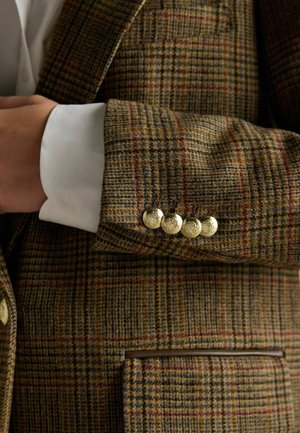 Brown and gold houndstooth blazer with gold embossed buttons, featuring textured fabric, a patch pocket, and a white shirt cuff visible.