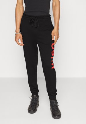 Persona que lleva pantalones deportivos negros con texto vertical rojo "HUGO" en la pierna izquierda, camiseta sin mangas negra, pulsera y zapatillas negras de caña alta.