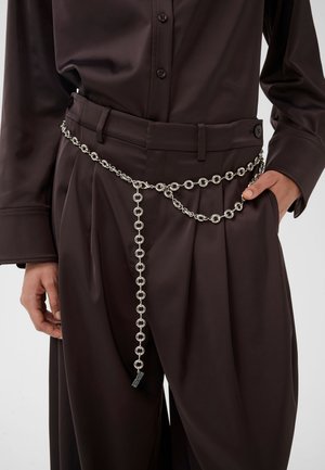 Persona vistiendo pantalones plisados de color marrón oscuro y camisa a juego, con un cinturón de cadena plateada que cuelga suelto en la cintura.