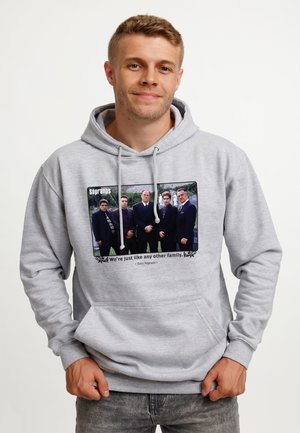Mężczyzna w jasnoszarej bluzie z kapturem z nadrukiem przedstawiającym pięciu mężczyzn w garniturach oraz tekstem "Sopranos" i cytatem Tony'ego Soprano.