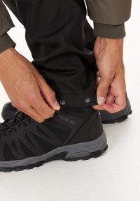 Händer som säkrar tryckknappar på svarta byxben över svarta och grå vandringsskor på vit golv.