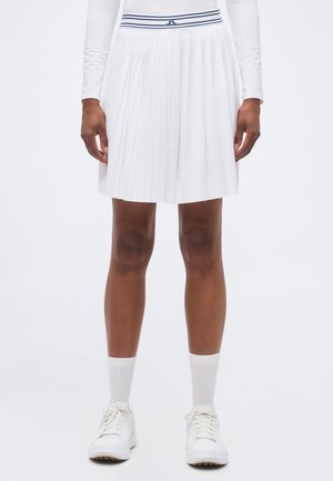 Mujer con una falda blanca plisada hasta la rodilla con cinturilla a rayas azules, camiseta blanca de manga larga, calcetines blancos y zapatillas blancas.