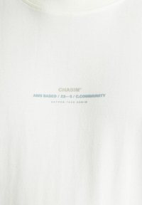 T-shirt bianco in cotone con scollatura rotonda e testo stampato in blu chiaro e grigio. Il design include dettagli sul marchio e sul prodotto.