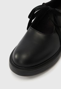 Μαύρο δερμάτινο παπούτσι με στρογγυλεμένη μύτη, επίπεδη λαστιχένια σόλα και μαύρα κορδόνια. Ο σχεδιασμός διαθέτει ομαλή υφή και ελάχιστες λεπτομέρειες.