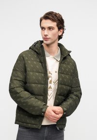 Maslinasto zelena puff jakna s kvadriranim texturom i suptilnim uzorkom, s kapuljačom i patentnim zatvaračem sprijeda, uparen s svijetlom košuljom.