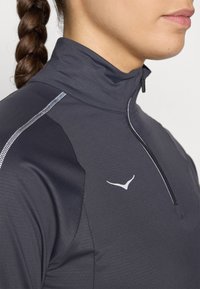 Pilkas sportinis megztinis su aukštu apykakle ir ketvirčio ilgio užtrauktuku, su atspindinčiomis detalėmis ir mažuoju baltu logotipu ant krūtinės.