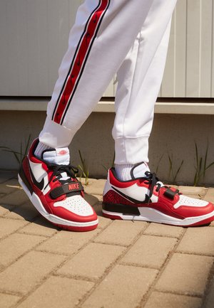 Røde og hvite Nike-sneakers med svarte detaljer; mønstret hæl, perforert tå og justerbar stropp. Brukes sammen med hvite joggebukser med rød stripe.