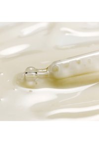 Lähikuva lasisesta tiputtimeen vapauttamassa kirkasta seerumin tippaa sileälle, kermaiselle valkoiselle pinnalle.