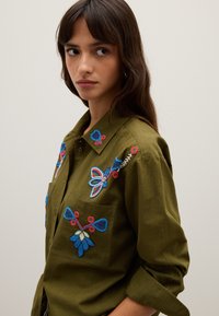 Jonge vrouw in een olijfgroene shirt met blauw en rood bloemmotief, kijkt opzij, tegen een effen lichte achtergrond.