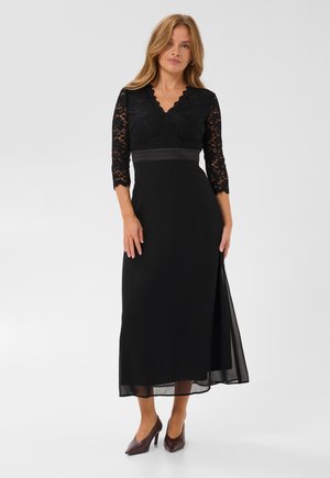 Femme portant une longue robe noire avec des manches en dentelle et un décolleté en V, debout devant un fond clair uni avec des talons hauts marron.