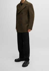 Olivgrön ull peacoat med dubbelknäppt design, stora svarta knappar och sidofickor, i kombination med svarta byxor och kraftiga stövlar.