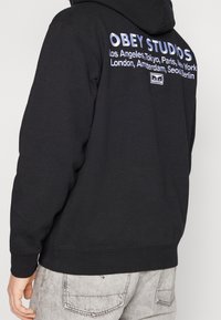 Černá mikina s kapucí s vyšitým nápisem „OBEY STUDIOS“ v bílé a stříbrné barvě na zádech, doplněná názvy měst. Měkká tkanina a standardní střih.