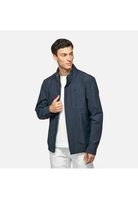 Marineblauwe lichtgewicht jas met een ritssluiting aan de voorkant, hoge kraag en zijzakken, met een gladde textuur en subtiele stikdetails.