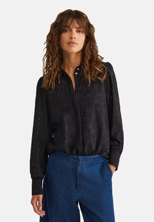 Blusa nera in pizzo con motivo floreale intricato, colletto con bottoni e maniche lunghe. Abbinata a pantaloni a gamba larga blu.