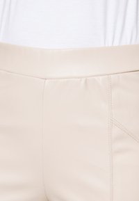 Pantaloni in faux leather beige con una superficie liscia, dotati di una vita piatta e dettagli cuciti lungo le cuciture laterali e frontali.