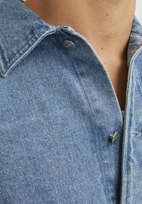 Denim košulja svijetloplave boje, sa šiljastim ovratnikom i srebrnim dugmadi na pritisak. Tekstura prikazuje klasični uzorak tkanja denima.