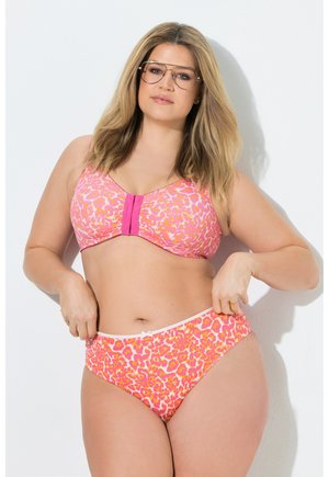 Femme grande taille portant un soutien-gorge rose et orange à imprimé léopard et une culotte assortie, ajustant la ceinture, sur un fond blanc uni.