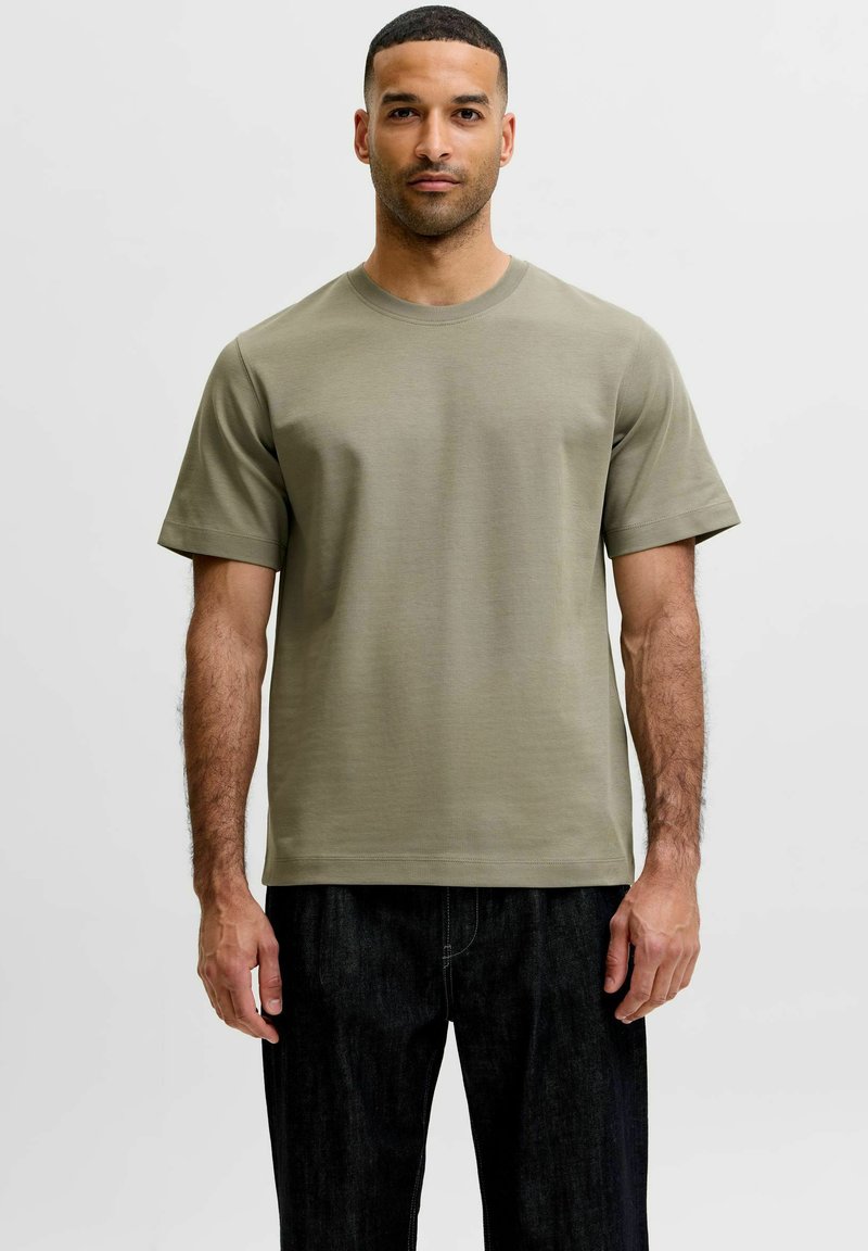 Άνδρας με ελιάς πράσινο κοντομάνικο T-shirt με στρογγυλή λαιμόκοψη και μαύρο παντελόνι, στέκεται με το πρόσωπο προς τα εμπρός σε απλό φωτεινό φόντο.