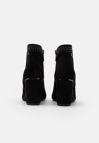 Botas de tobillo de ante negro con punta redonda, tacón alto grueso, detalles de tachuelas alrededor del tobillo y cierre de cremallera lateral.