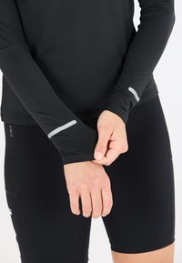 Zwarte atletische shirt met lange mouwen, een aansluitend ontwerp, vochtafvoerende stof en reflecterende accenten op de mouwen. De shorts zijn ook zwart en aansluitend.