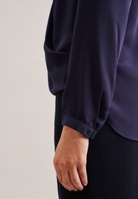 Blouse bleu marine avec une texture lisse, dotée de manches longues et de poignets boutonnés. L'ourlet est légèrement courbé pour un ajustement décontracté.