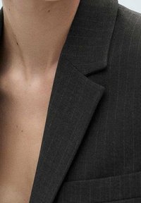 Tmavě šedé sako na míru se pruhovaným vzorem, výřezovým klopovým límcem a jednou náprsní kapsou. Látka má hladkou texturu a strukturovaný design.