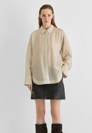 Kvinna med blött hår iförd en lös beige knappad skjorta, svart läderminikjol och svarta knähöga stövlar som står mot en enfärgad bakgrund.