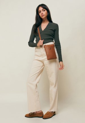 Žena oblečená v tmavozelenom top s dlhým rukávom, krémových nohaviciach s vysokým pásom, hnedých mokasínach, držiaca hnedú crossbody tašku na jednoduchom béžovom pozadí.