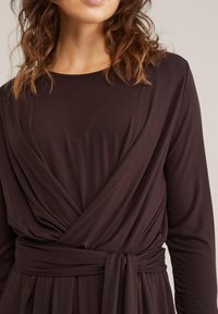 Vrouw draagt een donkerbruine jurk met lange mouwen en een wikkelriem in de taille, getoond van schouders tot midden torso tegen een neutrale achtergrond.
