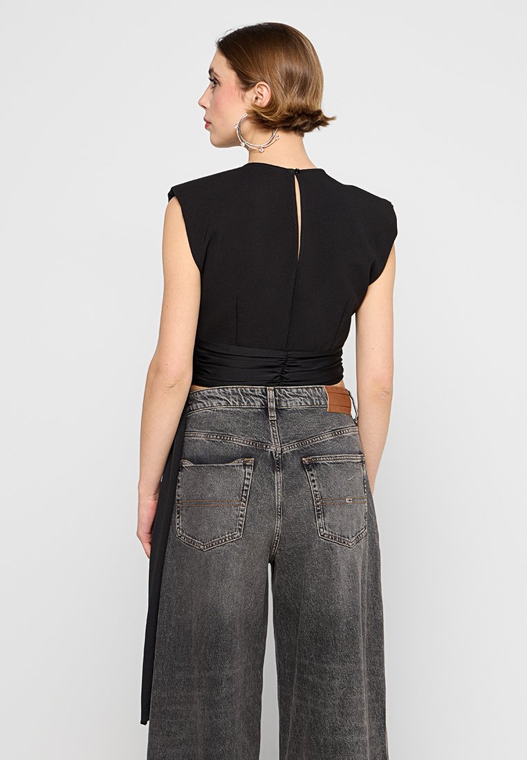 Frau mit kurzer Haarfrisur, die große Creolen trägt, ein ärmelloses schwarzes Oberteil mit einem Schlitz im Rücken und hochtaillierte, weite graue Jeans.