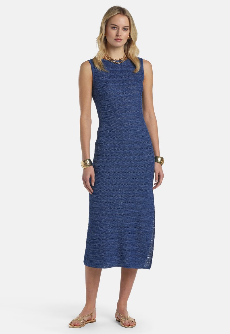 Femme portant une robe bleu tricotée, sans manches et longueur genou, avec des bijoux dorés et des sandales plates dorées, debout devant un fond blanc uni.