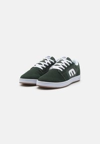 Tenisi verzi din suede, cu sireturi albe, talpă din cauciuc și un logo gri proeminent pe lateral, având un design plat și vârful rotunjit.