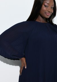 Granatowa, plisowana sukienka z długimi, luźno opadającymi rękawami i okrągłym dekoltem. Fakturowana tkanina dodaje elegancji; posiada lekko rozpięty przód.