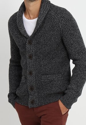 Homme portant un cardigan en maille gris foncé à col châle avec cinq boutons sur une chemise blanche et un pantalon brun rougeâtre, mains dans les poches.