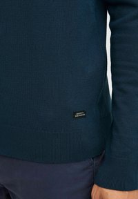 Námornícky modrý ľahký sveter vyrobený z mäkkého materiálu, s rebrovanými manžetami a lemom, na boku s malou čiernou nálepkou s nápisom "VENOVANÉ DOKONALOSTI DŽÍNSOV."