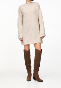 Abito maglione beige a maglia con maniche lunghe, scollo rotondo, vestibilità ampia e texture a coste, abbinato a stivali alti fino al ginocchio in suede marrone.