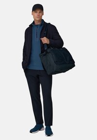 Lässiges marinefarbenes Sportoutfit mit einem dunkelblauen Oberteil mit Reißverschluss, dunkler Hose und blauen Turnschuhen, ergänzt durch eine dunkle Sporttasche und eine Kappe.