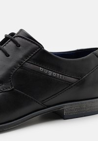 Chaussure en cuir noir avec une surface texturée, un design minimaliste, des coutures contrastées et un logo discret sur le côté.