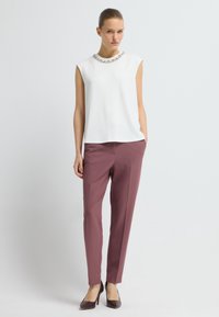 Frau in einem ärmellosen weißen Oberteil mit verziertem Ausschnitt, mauve-farbigen, taillierten Hosen und dunkelbraunen High Heels, die mit Händen in den Taschen steht.