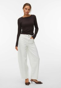 Femme portant un haut noir moulant à manches longues, un pantalon blanc large taille haute, et des talons bruns pointus ornés de nœuds et de points blancs.