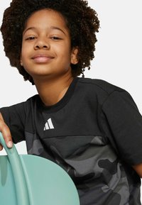 Black t-shirt with a camo design, featuring a white logo on the chest. The child is seated, holding a mint-colored chair. Curly hair visible.