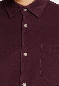 Närbild av en mörk lila kordöljskjorta med krage, fyra knappar och en bröstficka på höger sida.