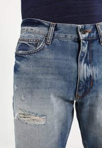Light blue denim jeans with a faded finish, featuring a distressed patch and a frayed rip on the thigh, standard five-pocket design.