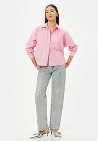 Camicia a maniche lunghe rosa con bottoni e taschino sul petto, abbinata a jeans dritti in denim chiaro e mule argentate con tacco.