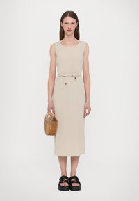 Rochie tricotată bej cu un design ajustat, talie strânsă cu un cordon și o lungime până la genunchi. Asortată cu un coș din lemn și sandale cu platformă.