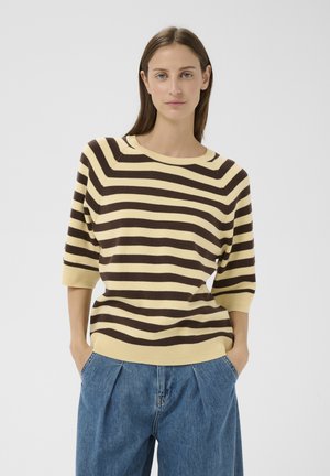 Egyenes, barna hajú nő bézs és sötétbarna csíkos pulóvert és bő kék farmert visel, kezét zsebében tartva áll.