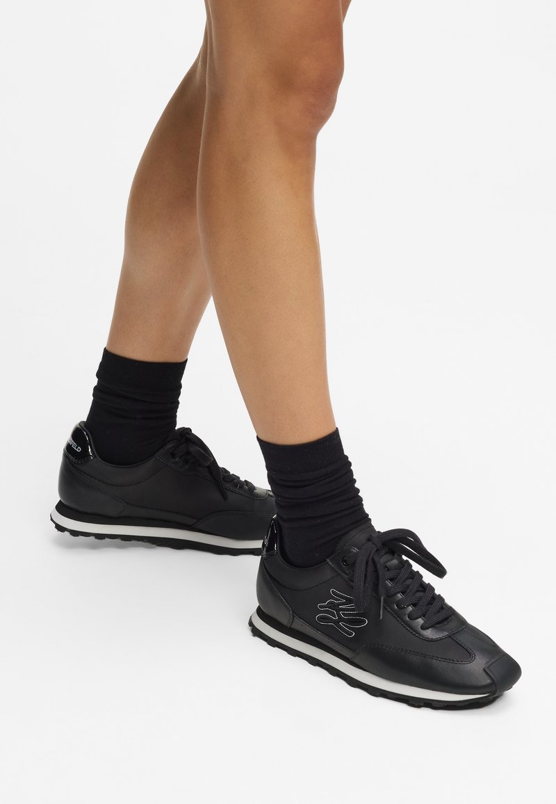 Baskets de sport noires avec une tige en cuir lisse, semelle en caoutchouc blanche et lacets noirs. Présente un design latéral texturé et un col rembourré.