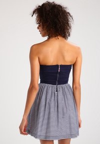 Kvinna med lockigt hår som bär en ärmlös marinblå topp och kjol i vitt med marinblå horisontella ränder, vänd bort med dragkedjan synlig på ryggen.