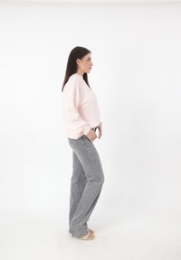Helles Rosa, übergroßer Pullover mit umgeschlagenen Ärmeln, kombiniert mit grauen Jeans in hoher Taille. Das Model steht im Profil und trägt beige Sandalen.