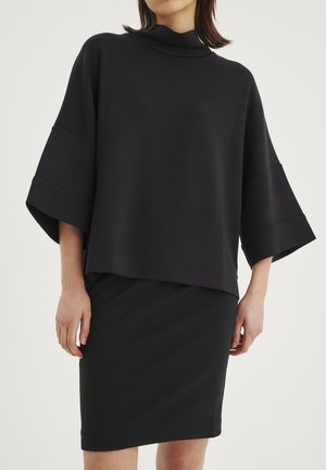Top nero oversize con collo alto e maniche ampie, abbinato a una gonna nera aderente. Tessuto morbido con una texture liscia, design minimale.